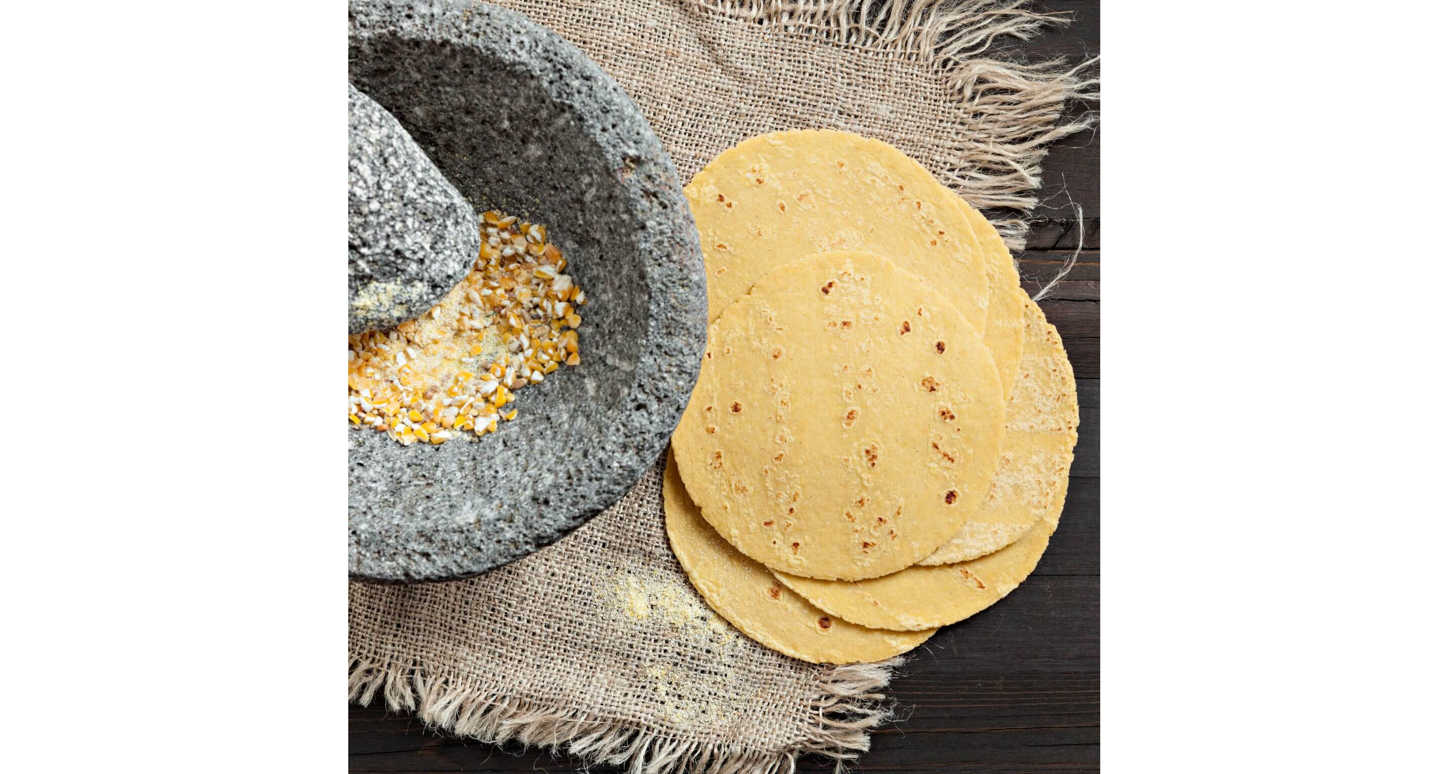 A wooden table with homemade tortillas next to a stone grinder filled with dried corn.