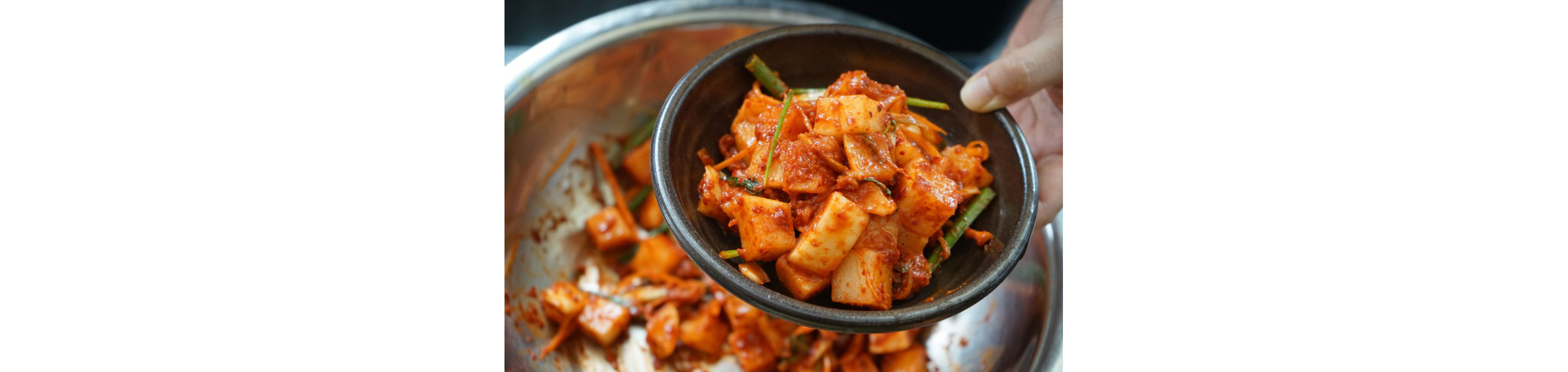 A person holding a small bowl filled with kimchi from a larger, stainless steel bowl.