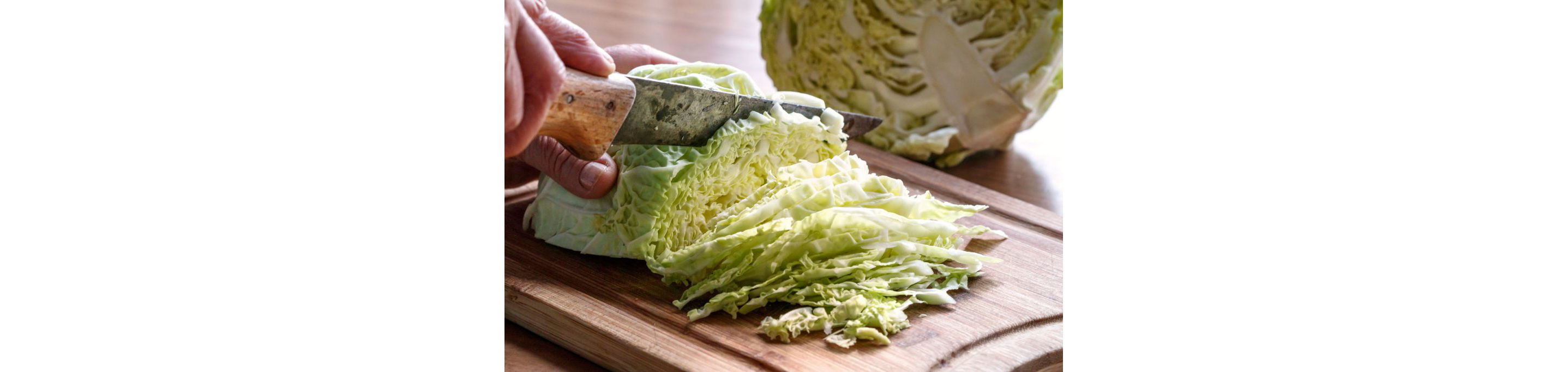 A person thinly chopping a halved cabbage on a wooden cutting board.