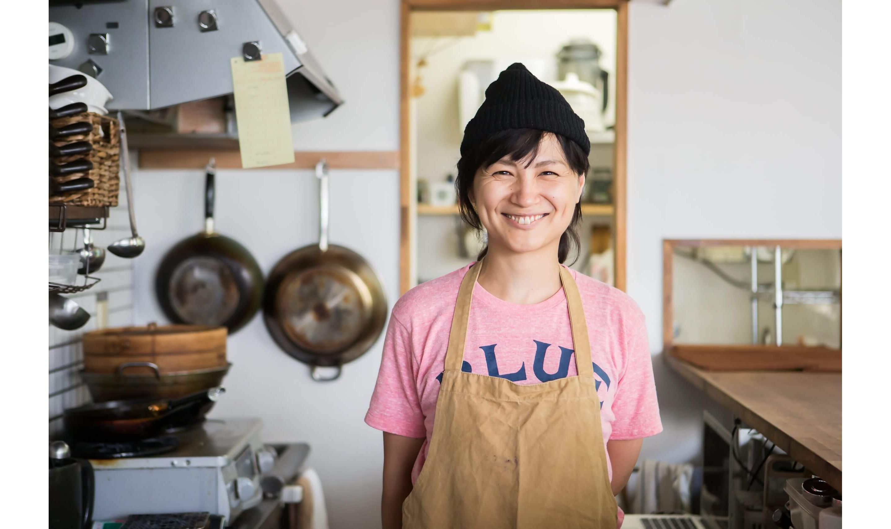 A woman in a canvas apron and pink shirt happily smiling at you from her kitchen.