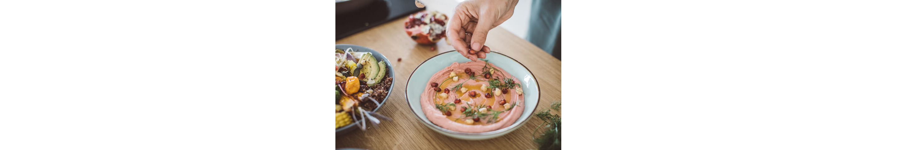 A soft hand sprinkling pomegranate on smooth hummus with herbs and whole chickpeas.