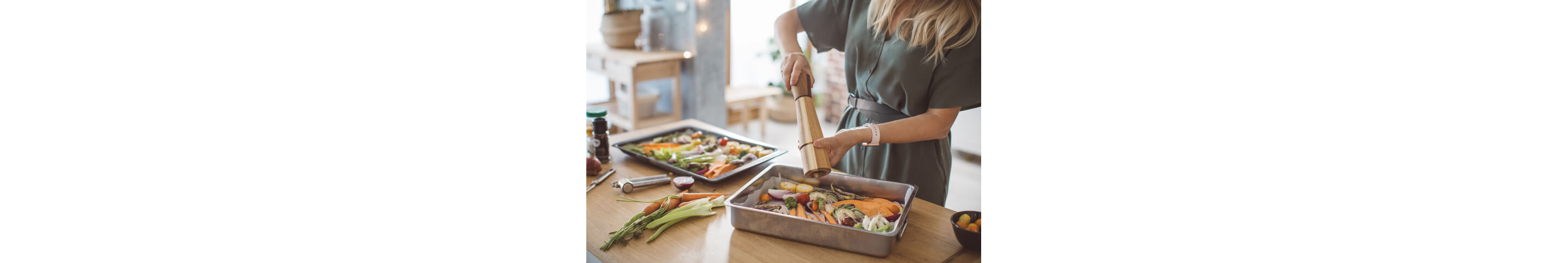 A woman seasoning chopped vegetables in a baking tray with a wooden pepper grinder.
