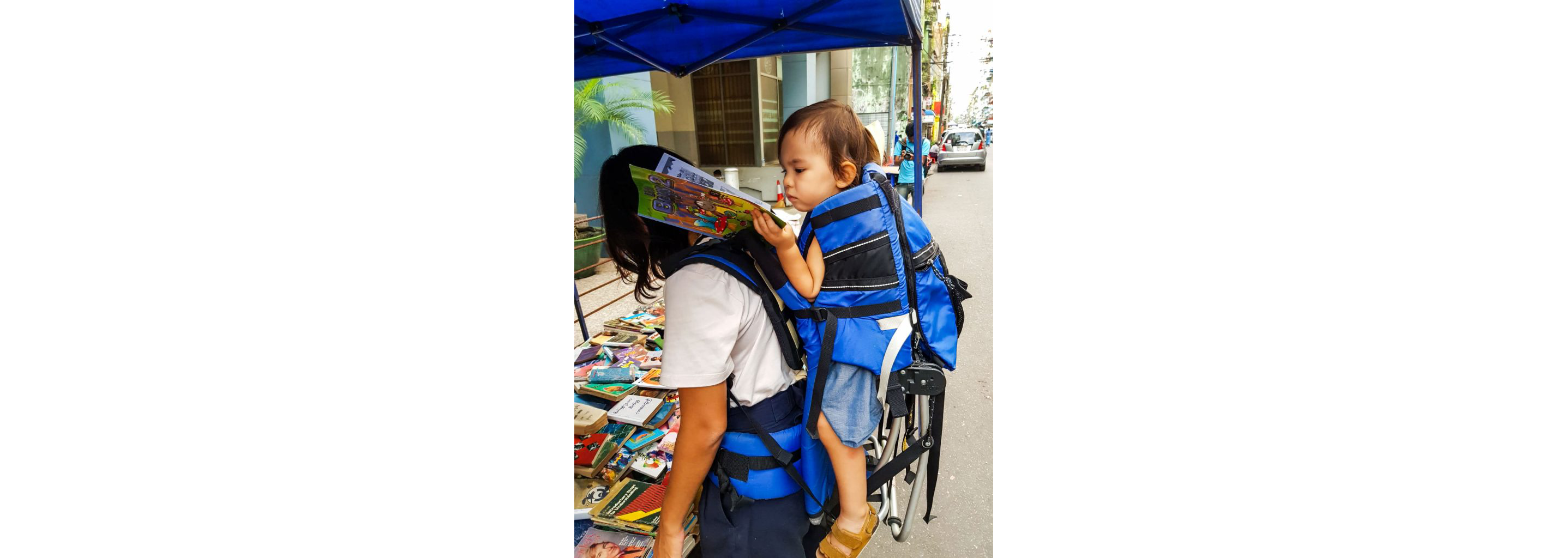 Wee Ling Soh's daughter reading a magazine while being carried in a blue backpack carrier.