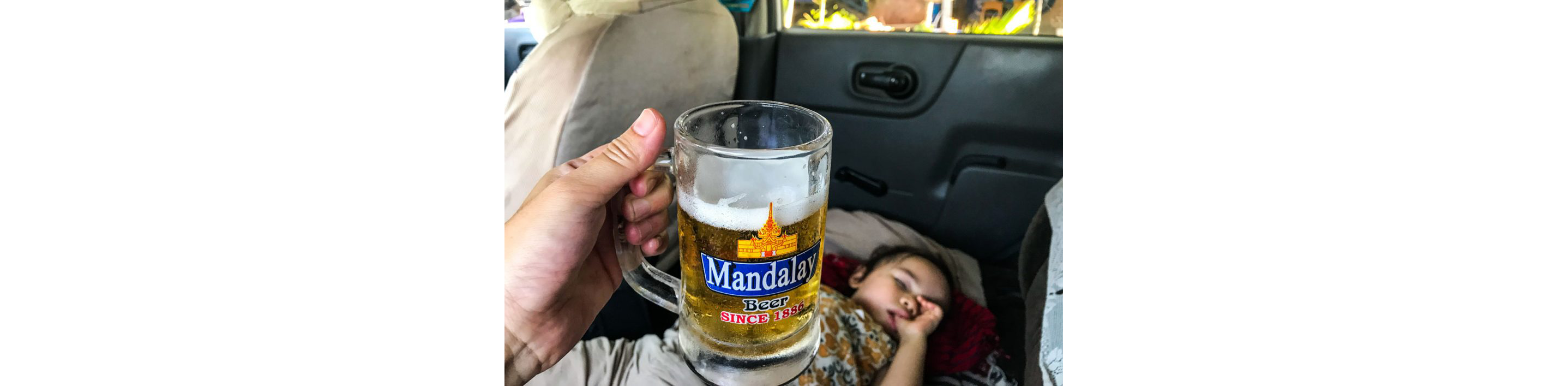 A hand holding a local lager in the back of a car with a sleeping child.