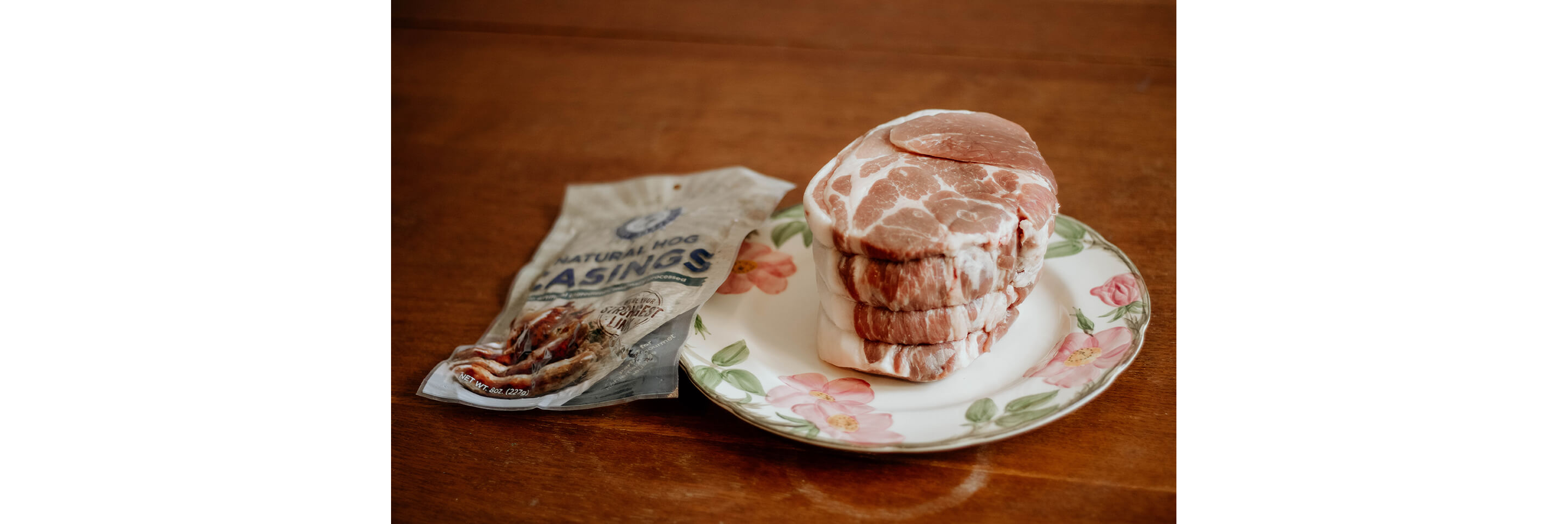 Meat resting on a floral china plate next to an unopened bag of natural hog casings.