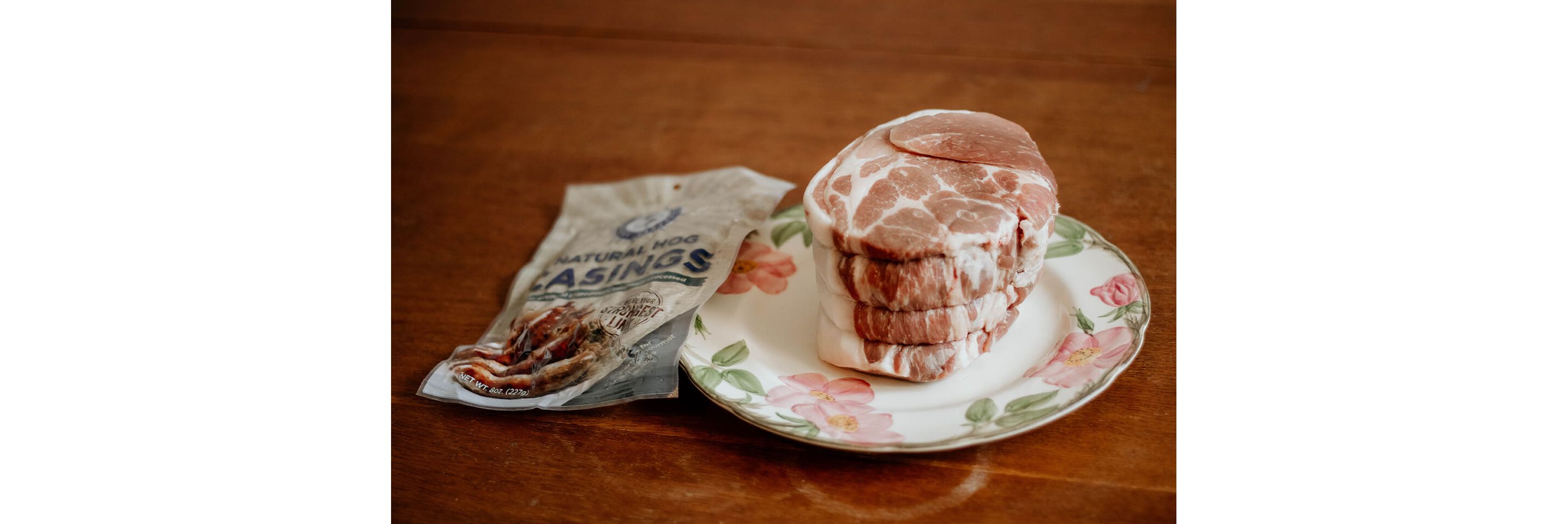 Meat resting on a floral china plate next to an unopened bag of natural hog casings.