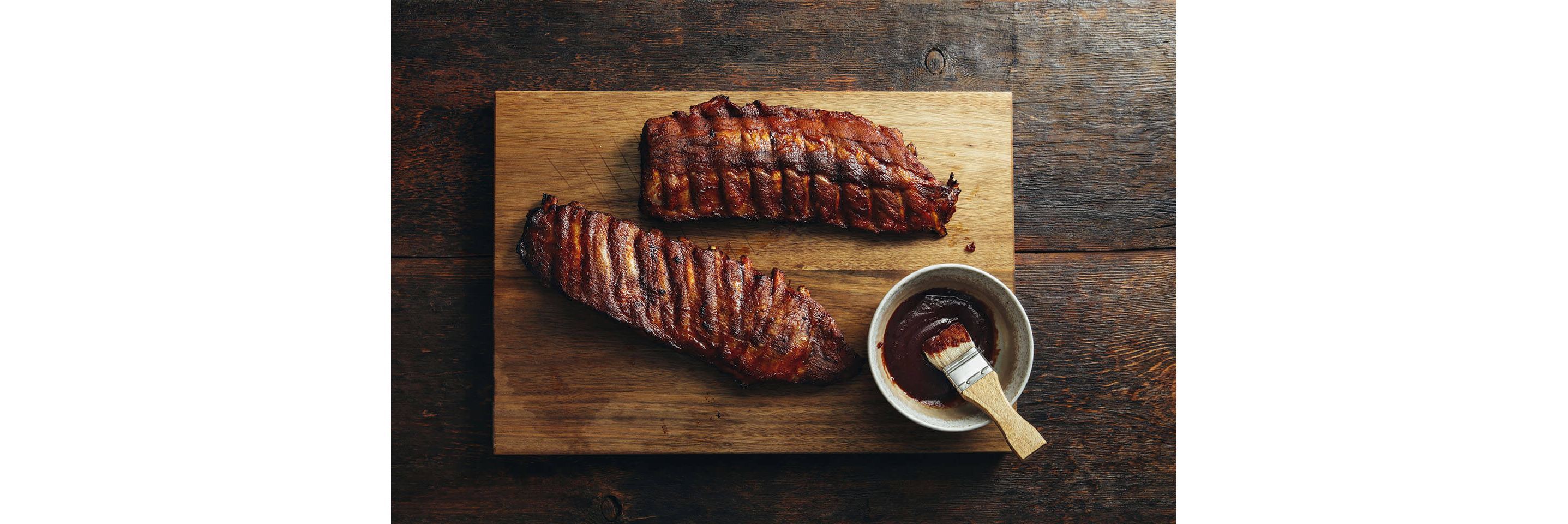 A rack of ribs resting on a wooden cutting board next to a bowl of barbeque sauce.