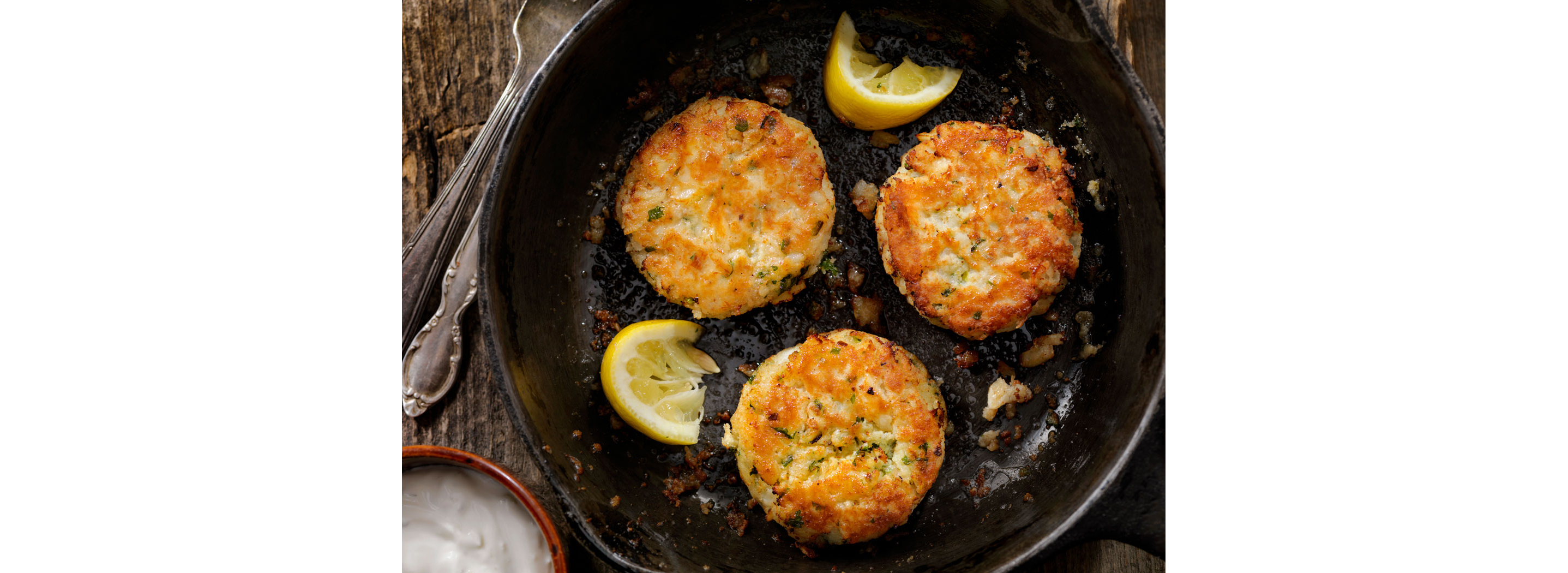 Three crab cakes frying in a skillet with sliced lemon.