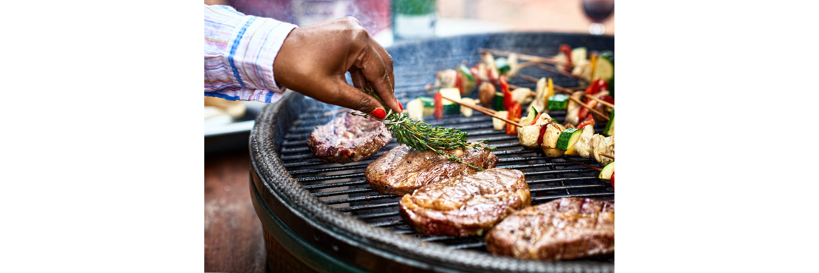 A hand basting grilled steaks in olive oil with a tied bunch of thyme.