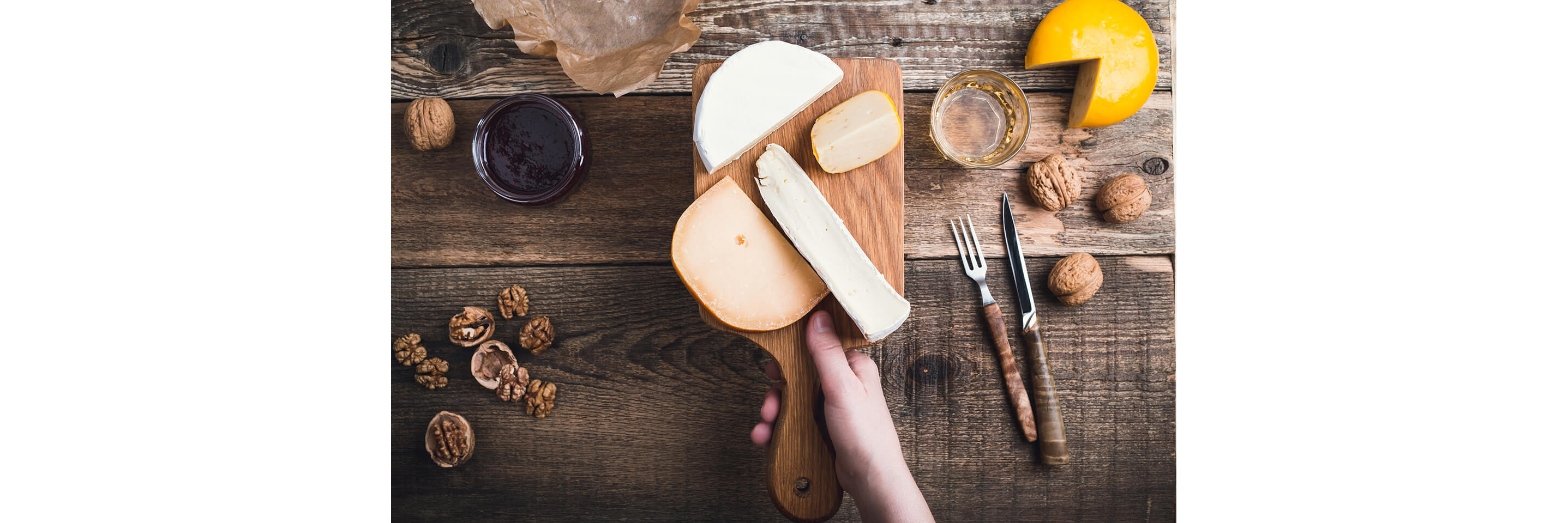 A hand holding up a rustic, wooden cheese board with assorted cheeses.