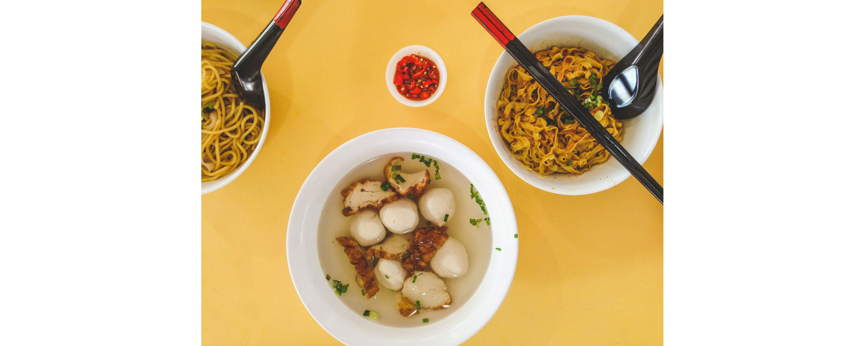 Bowls of Singaporean noodles and soup on a bright yellow table.