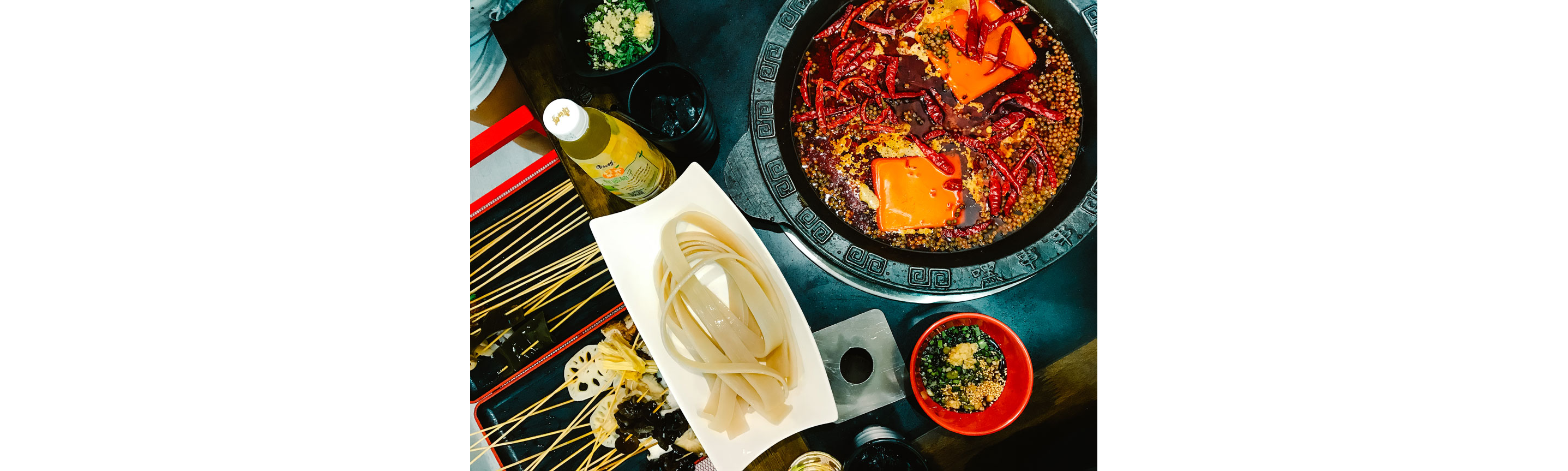 A white dish holding noodles next to a spicy steamboat.