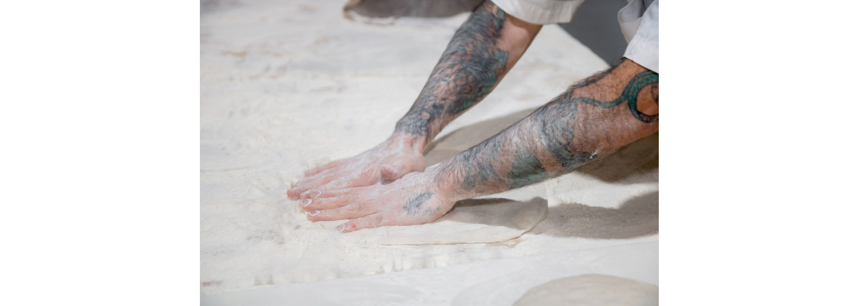 A person flattening dough surrounded by flour.