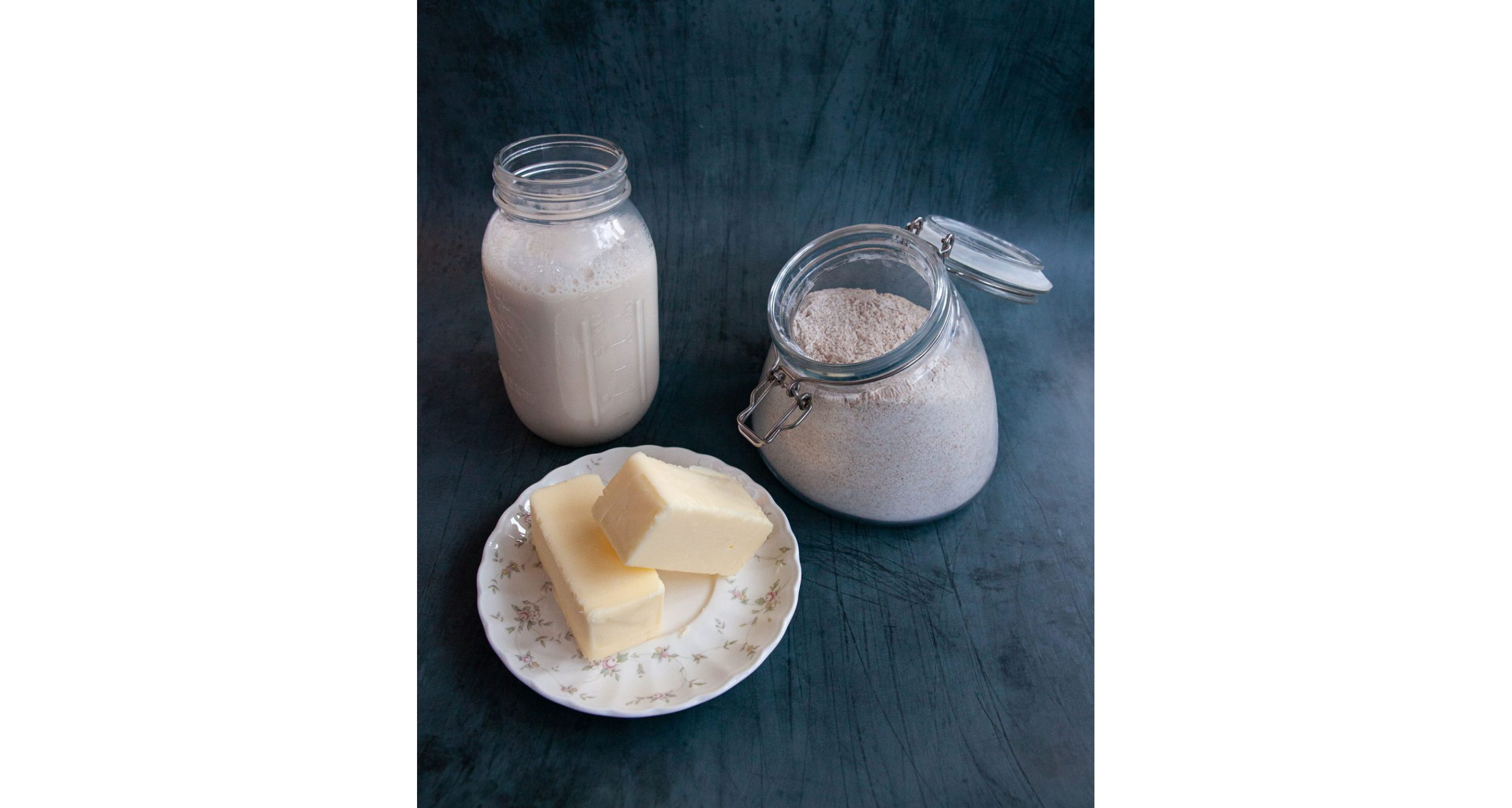 A small jar of milk, a plate of butter and a container of flour resting on a table.
