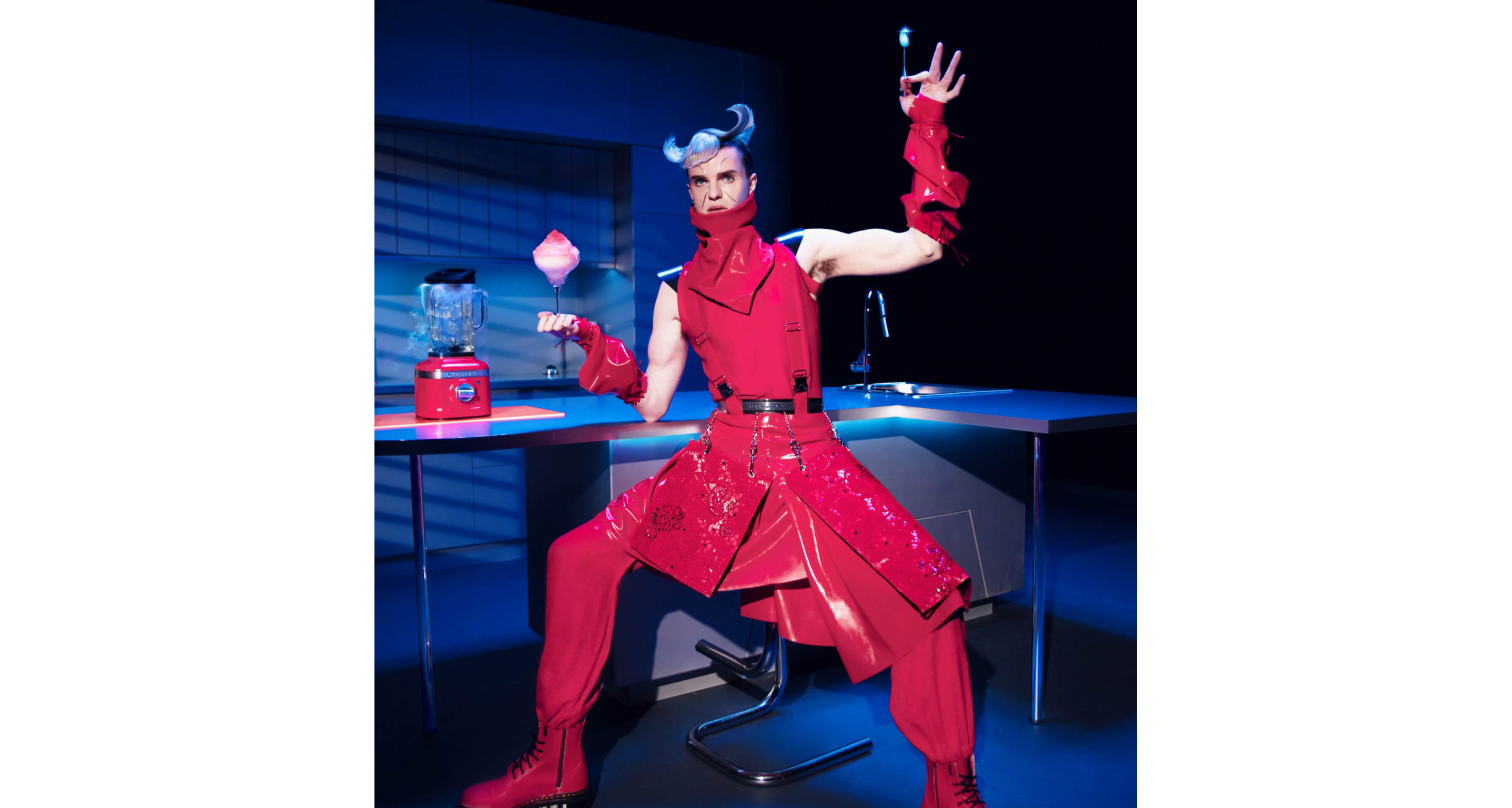 A model posing in a kitchen wearing Look Five while holding a glass filled with whipped pink cream near the K400 Blender in Hibiscus. 