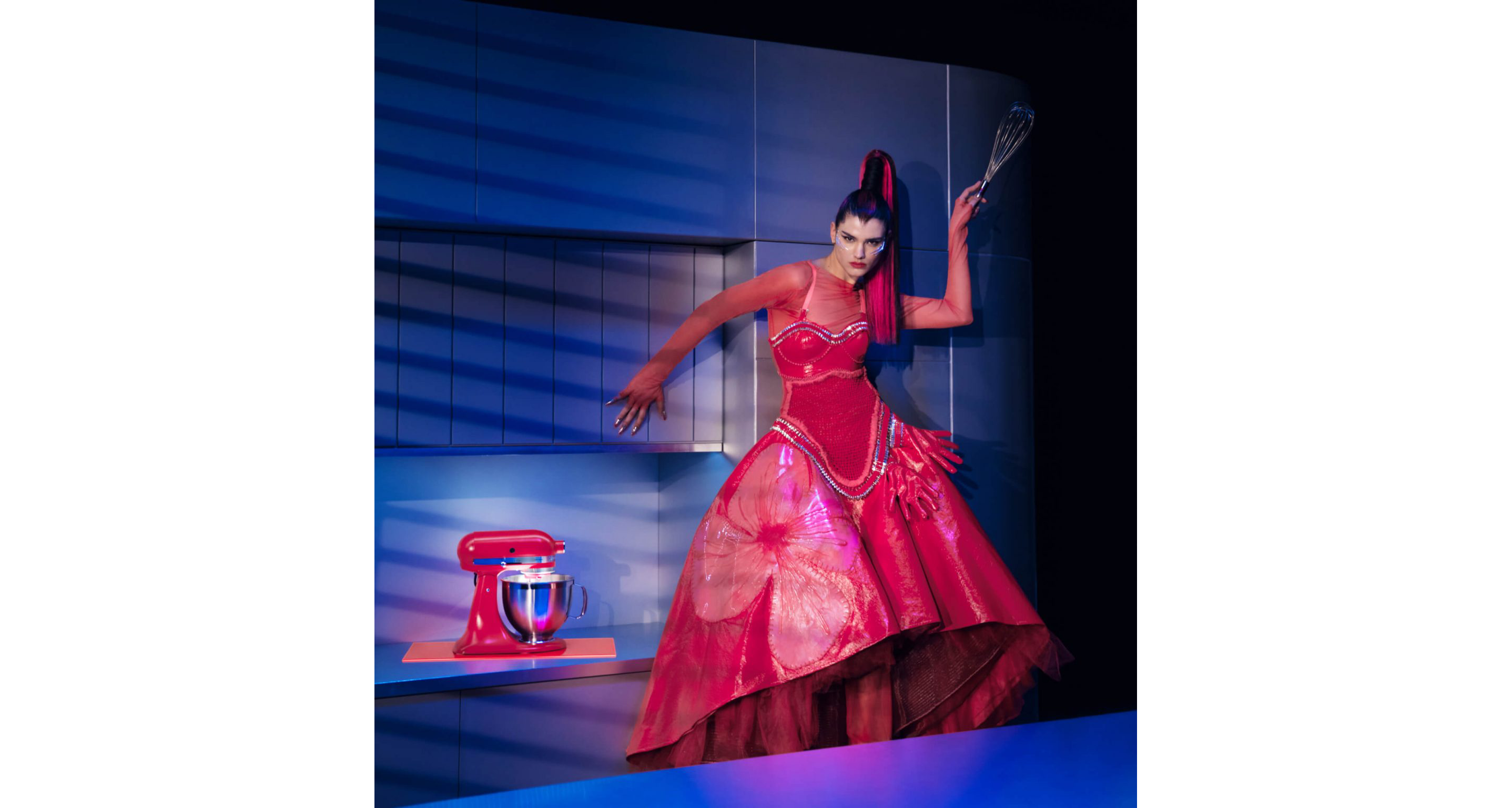 A model posing in a kitchen wearing Look Nine while holding a whisk next to the Artisan® Series Stand Mixer in Hibiscus on the countertop. 