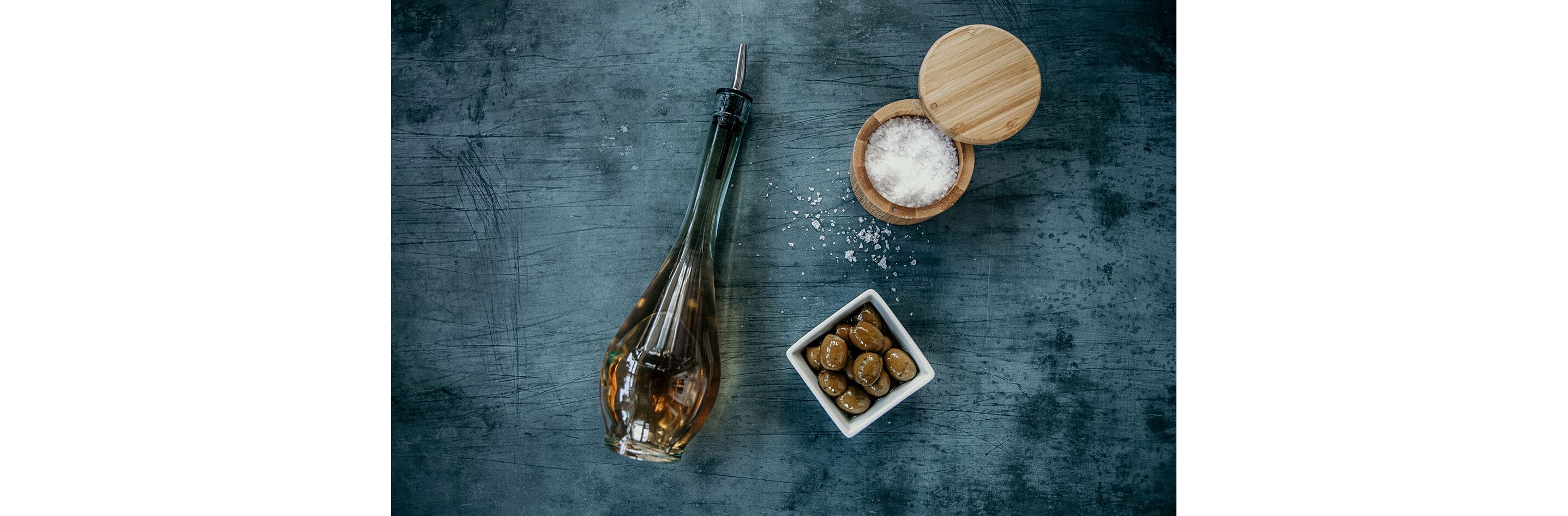 A sleek, skinny bottle of vinegar next to a dish of olives and a wooden container of kosher salt.