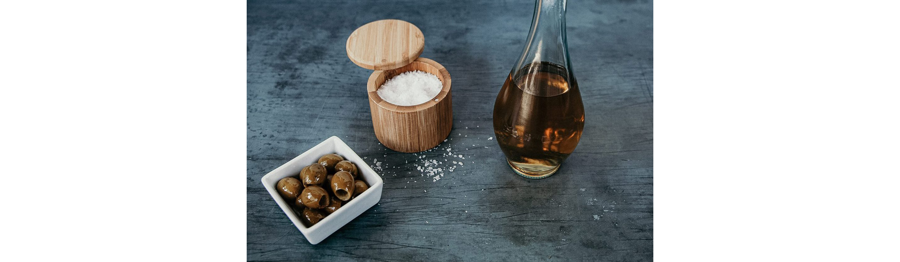 A bottle of vinegar, a dish of olives and a container of kosher salt resting on a table.