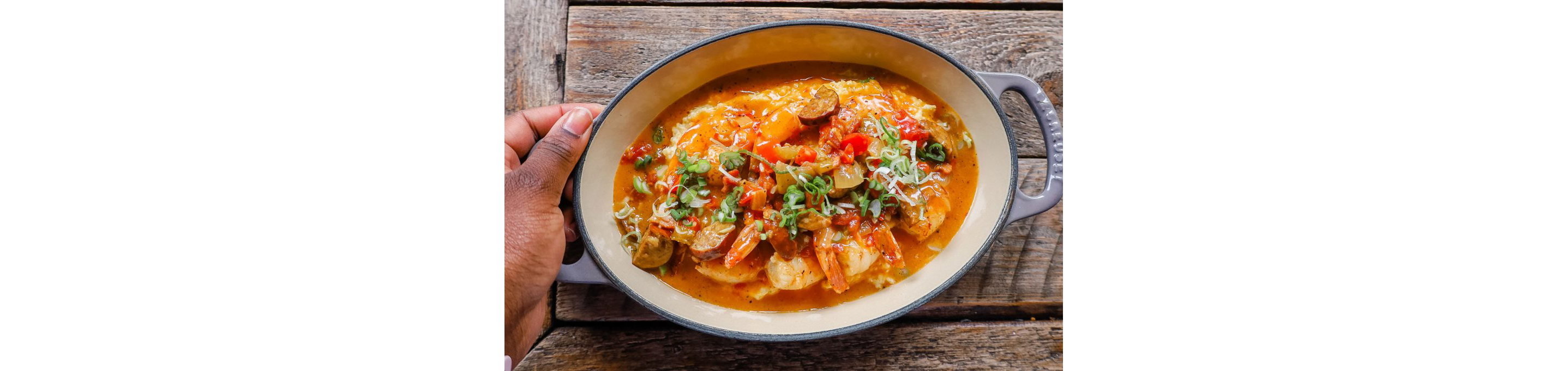 A person holding an oval dish filled with steaming cajun shrimp and grits.