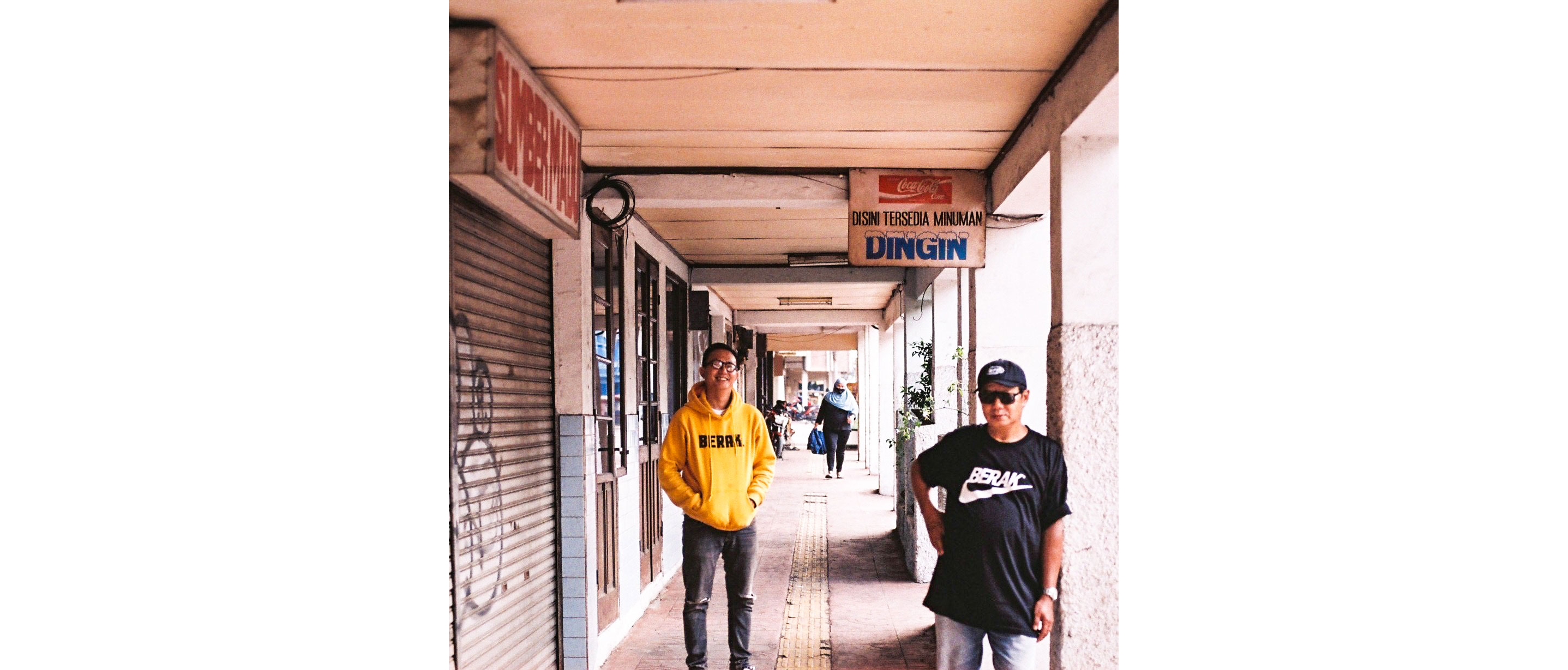 Paulus Supomo in black sunglasses leaning against a cement wall with son Yulius Iskandar in yellow sweatshirt standing behind.