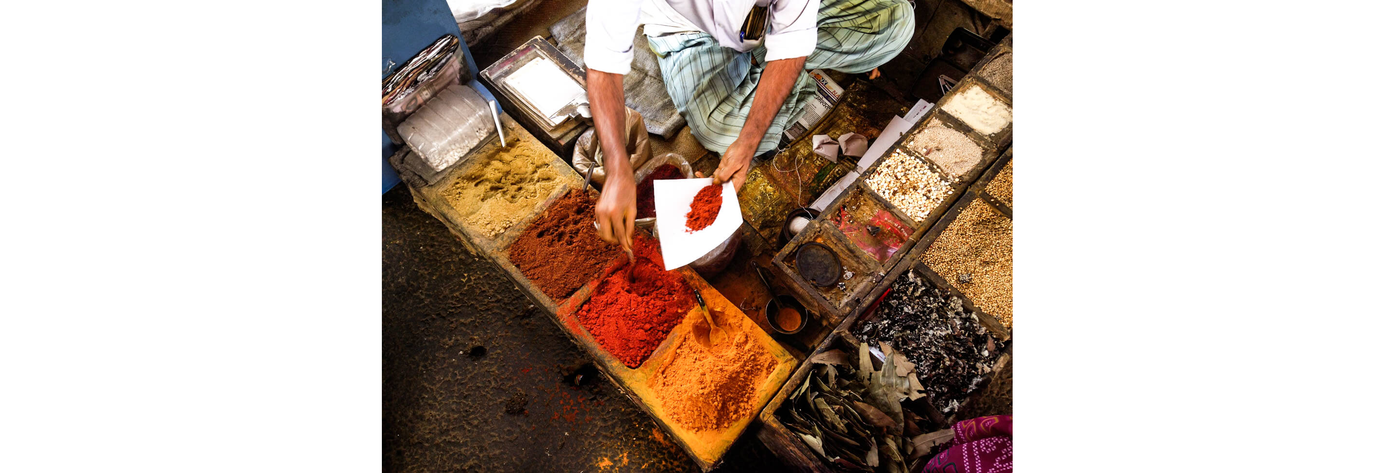 A person collecting a red spice amongst a variety of colorful spices, seasonings and dried herbs.