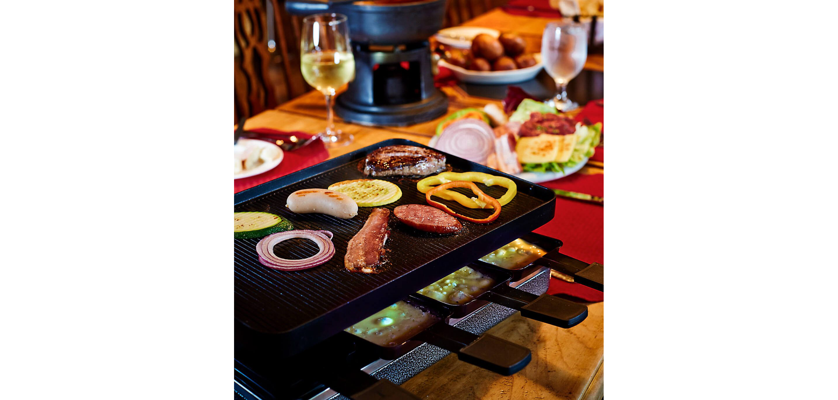 A raclette grill with a griddle on top cooking various meats and vegetables.