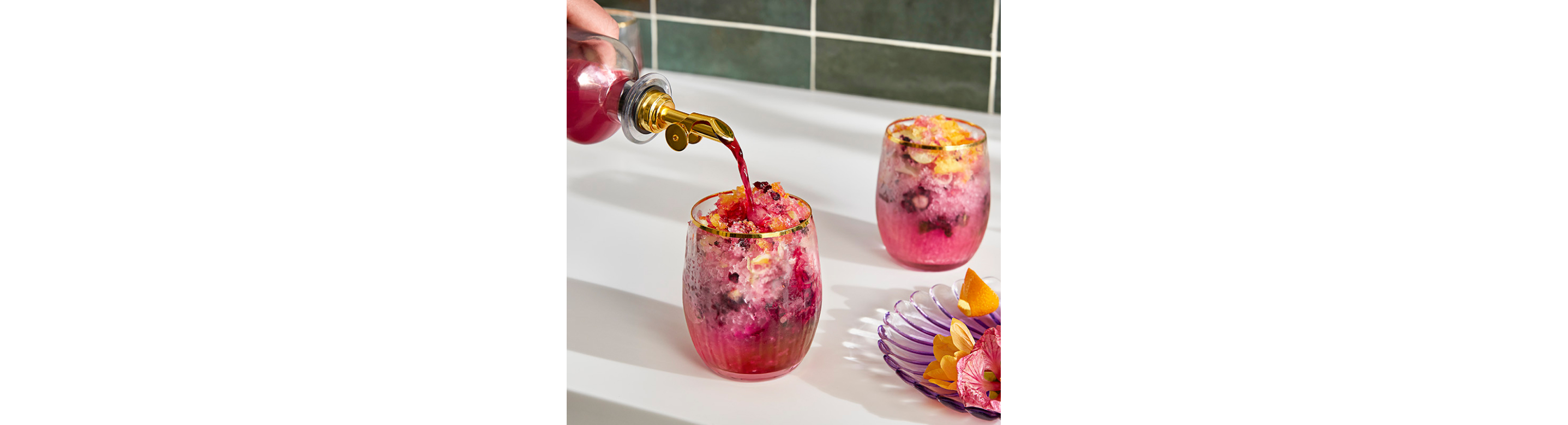 A person pouring hibiscus syrup into a pink shaved ice drink with floral garnishes.
