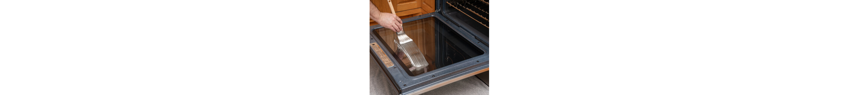 The inside of oven door glass with a hand painting a cleaning paste on it in the foreground