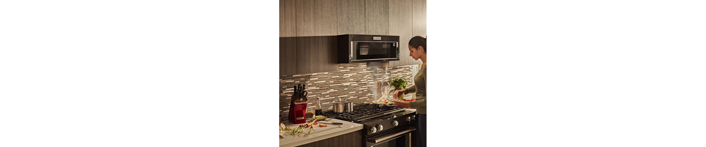 Person preparing stovetop recipe while food heats in a KitchenAid® low profile microwave above