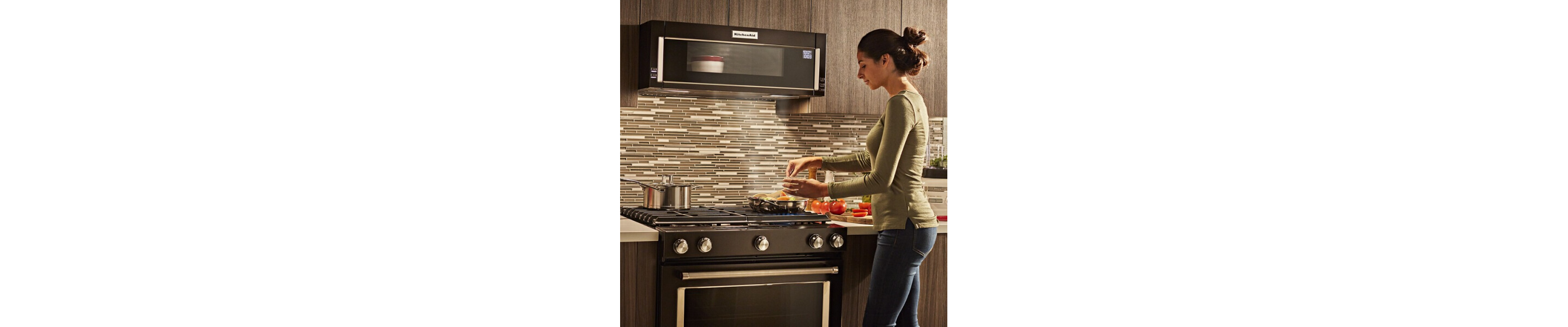 Person cooking food on a gas stovetop below a KitchenAid® low profile microwave warming food