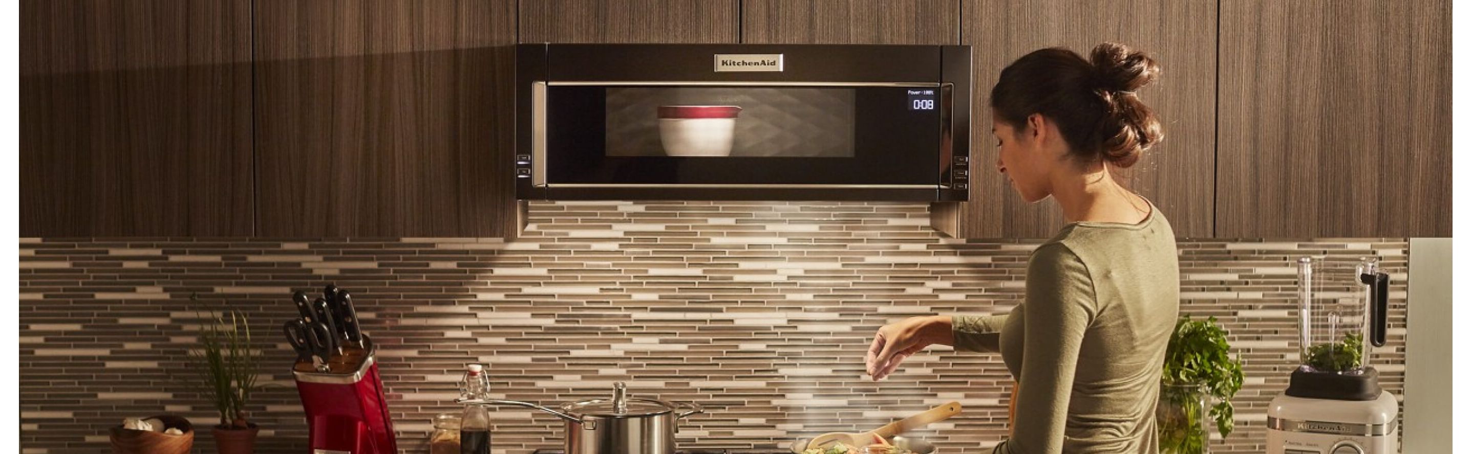 Person preparing a recipe on a gas stovetop below a KitchenAid® low profile microwave