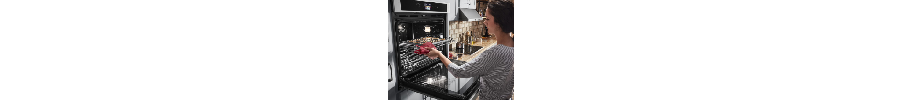 A woman pulling a baking tray out of a wall oven with a read potholder