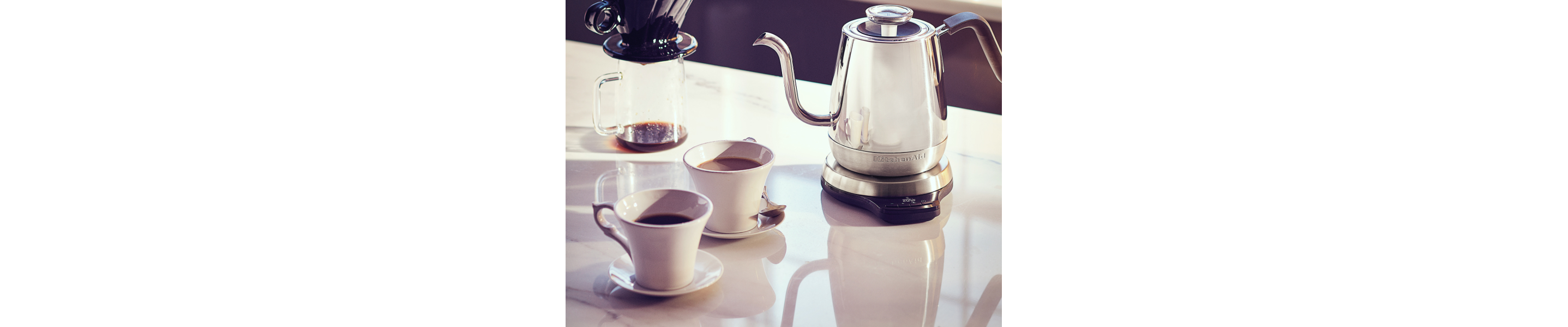 Silver KitchenAid® gooseneck electric kettle on white countertop with pour over coffee container and two cups of coffee