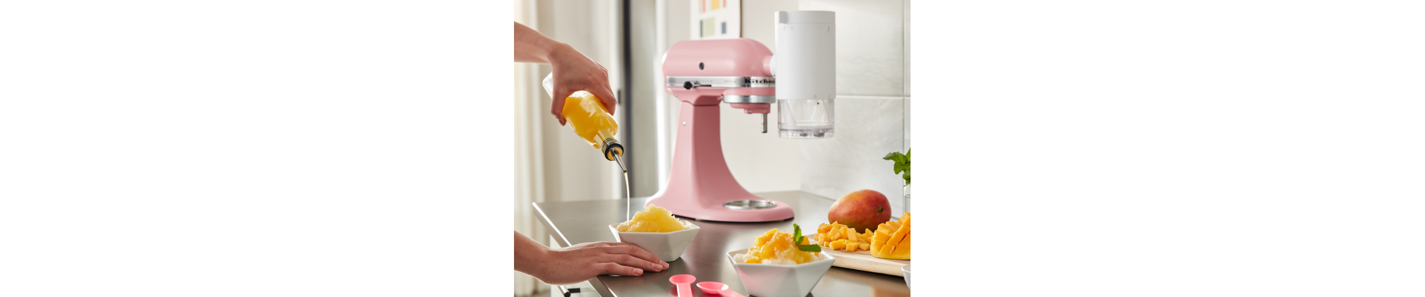 Person pouring syrup over shaved ice next to a KitchenAid® Shave Ice Attachment on a pink KitchenAid® stand mixer 