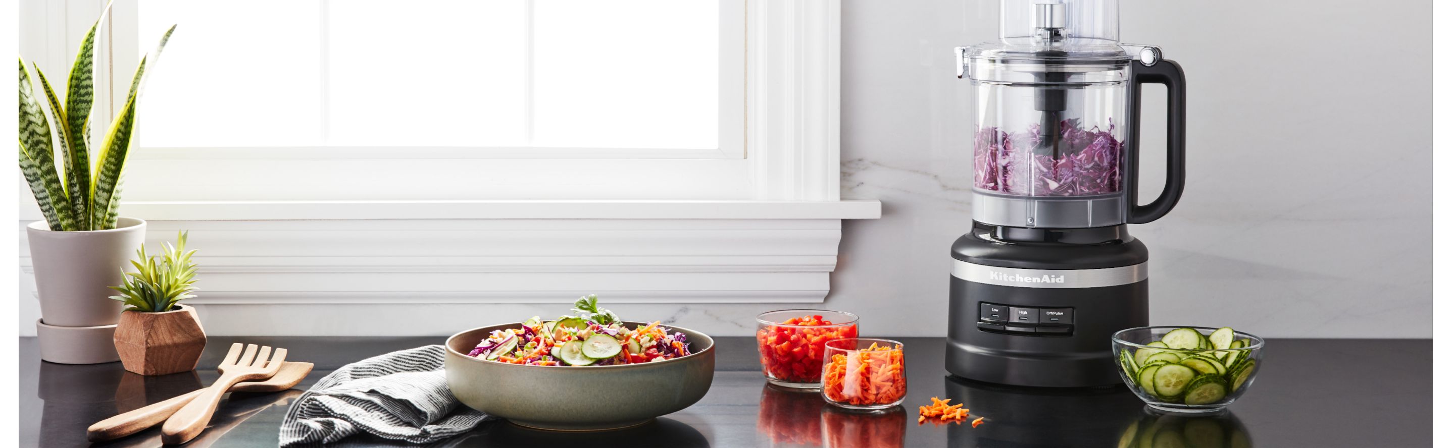 Black KitchenAid® food processor full of shredded red onion next to prepared veggies on the countertop