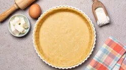 A tart crust and various baking implements and ingredients on a grey countertop.