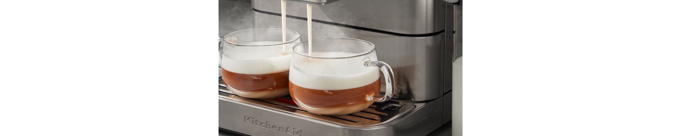 A pair of glass espresso cups filled with espresso, with steamed milk pouring into the glasses from the machine's dual spouts.