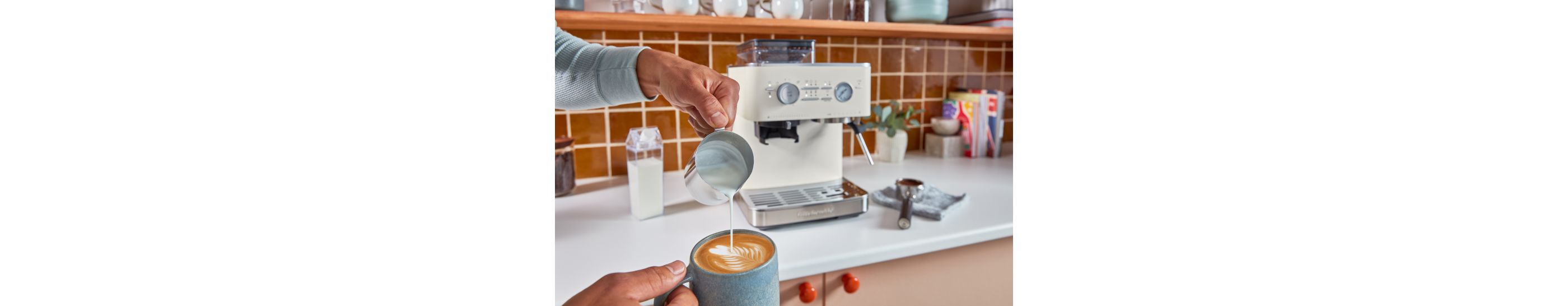 A person pouring steamed milk into a cup of espresso with a KitchenAid® Espresso machine in the background.