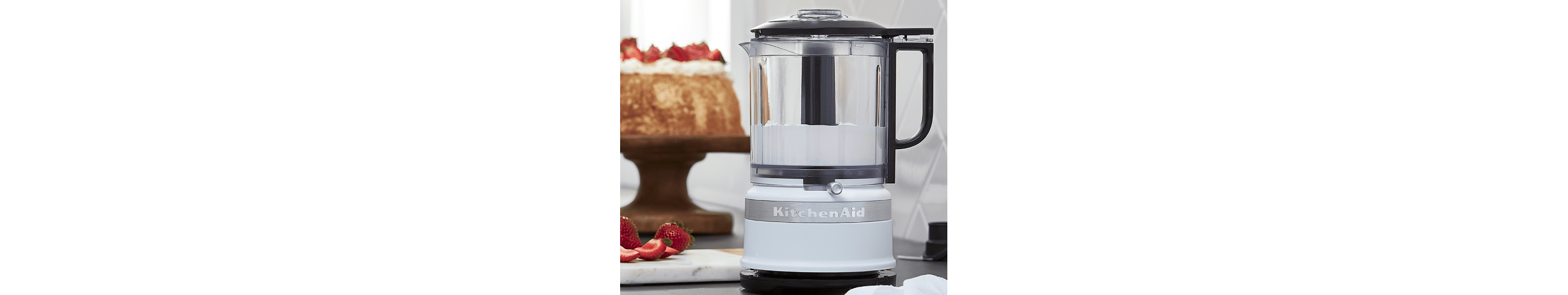 A KitchenAid® food processor in front of a cake topped with strawberries