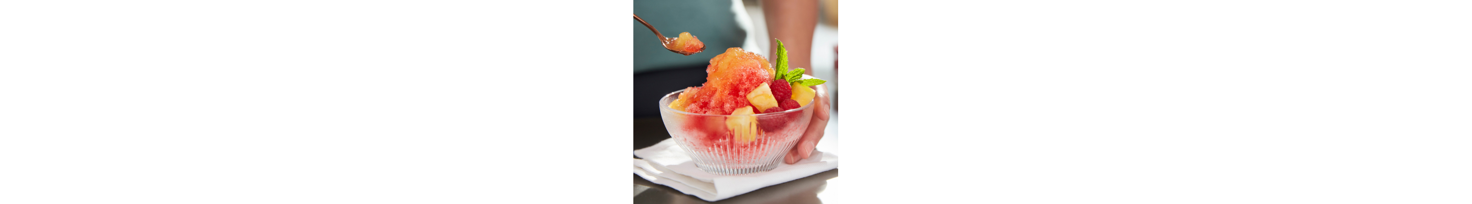 Person placing spoon in shave ice dessert topped with chopped fruit and syrup