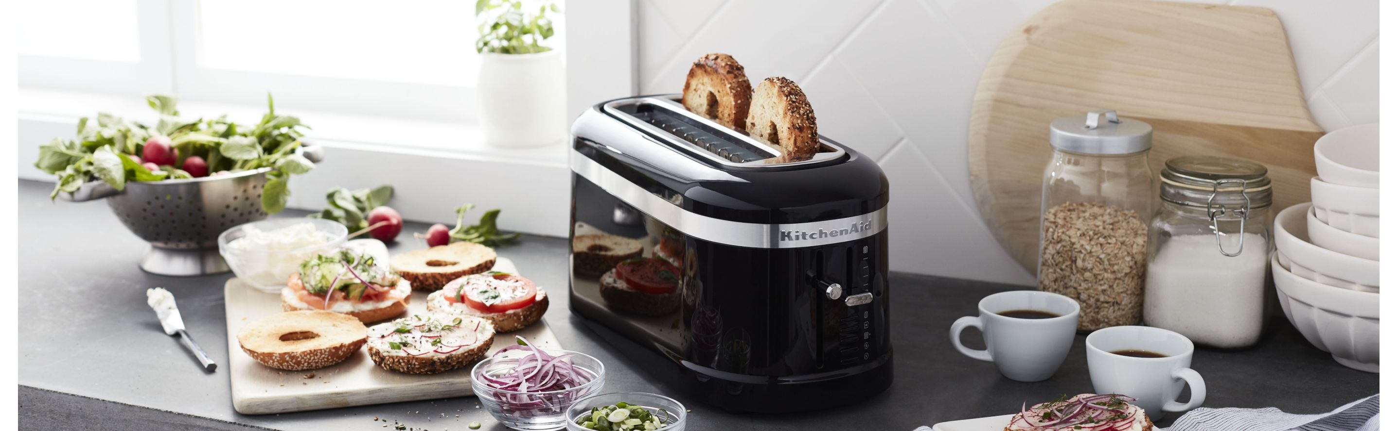 Bagel slices in a pop-up KitchenAid® toaster on a modern kitchen counter next to bagel sandwich fixings.