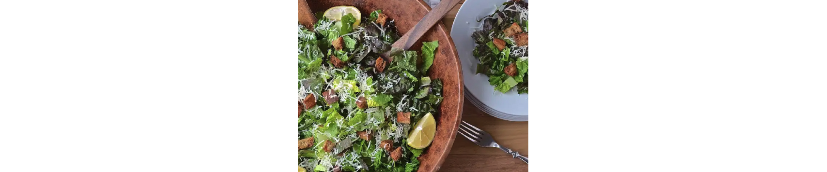A wooden serving bowl with chopped salad next to a place setting with salad.