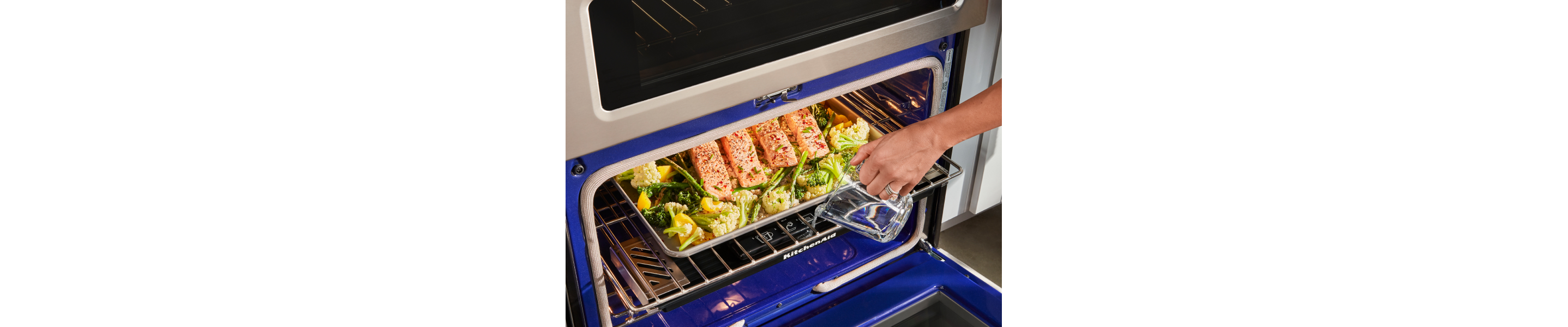 A woman checking on salmon and vegetables on a baking sheet in a KitchenAid® oven.