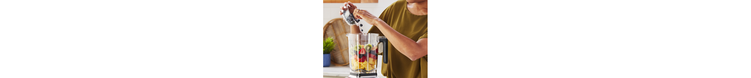 A woman adding blueberries to a KitchenAid® blender that has a variety of fruit