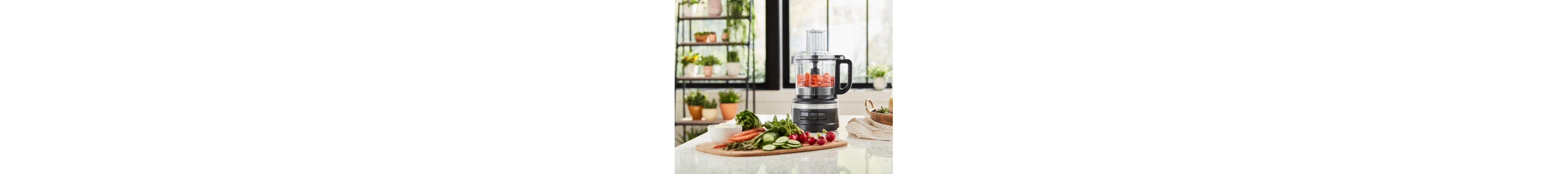 Black food processor filled with sliced carrots next to various sliced vegetables
