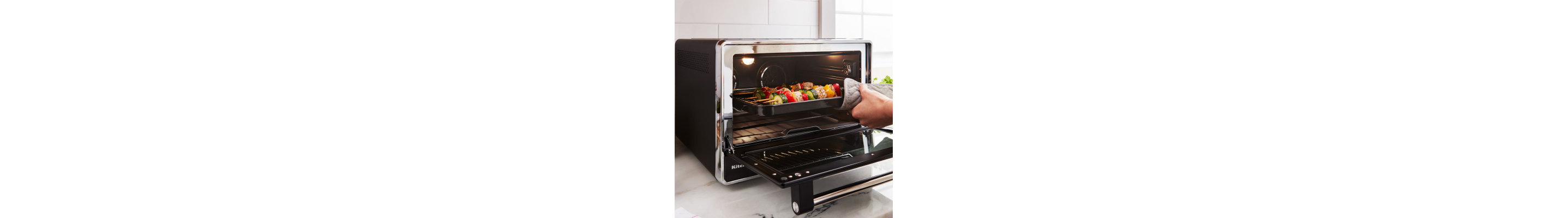A person holding a tray of kebabs cooking inside a black countertop oven