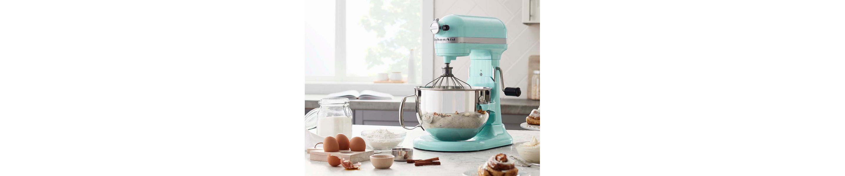 KitchenAid® stand mixer with a wire whip accessory next to cracked egg shells and fresh baked cinnamon rolls.