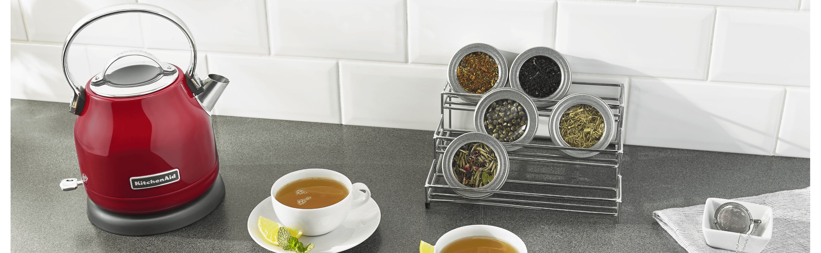 KitchenAid® electric tea kettle on countertop with cups of tea and containers of loose tea leaves.