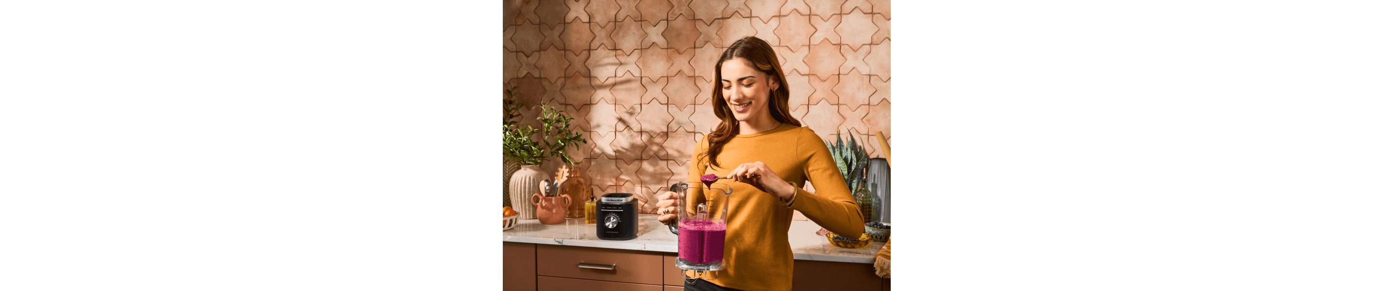 Woman with berry smoothie in a blender standing in front of wooden cabinetry and a white marble countertop