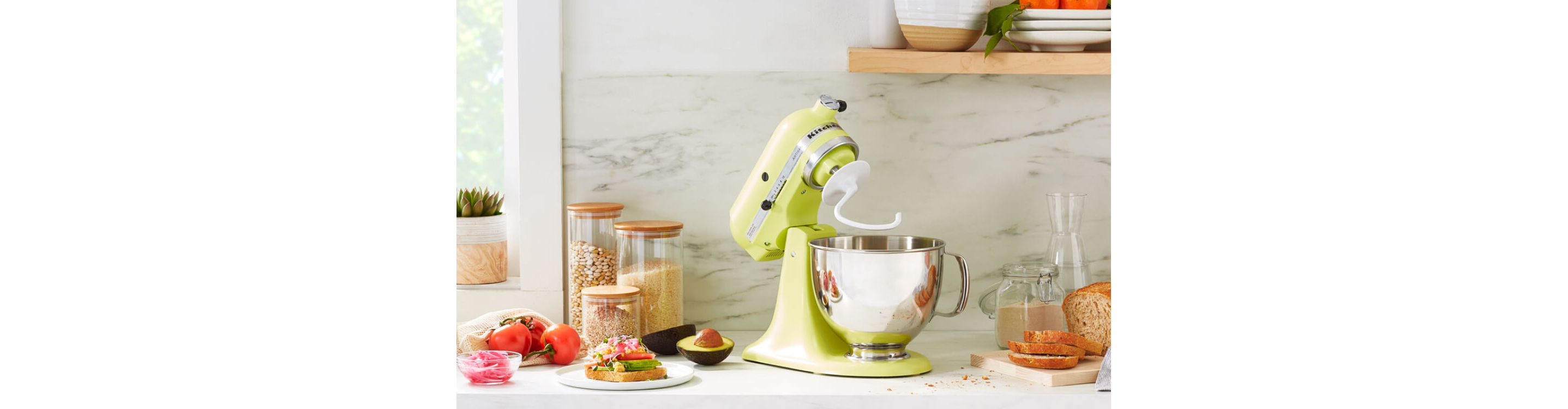A KitchenAid® stand mixer with a dough hook accessory on a modern kitchen counter next to fresh vegetables for making a sandwich.
